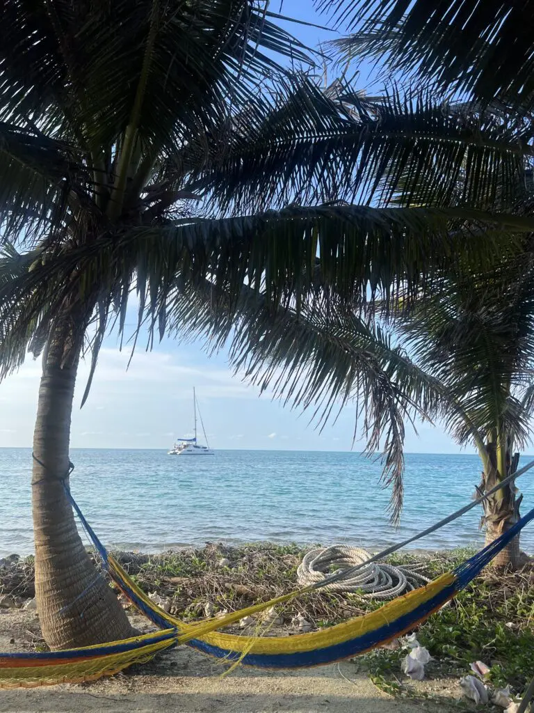 A picturesque hammock on the beach, perfectly positioned to take in the breathtaking view of a sailboat gliding across the horizon during your unforgettable Belize Sailing Vacation.