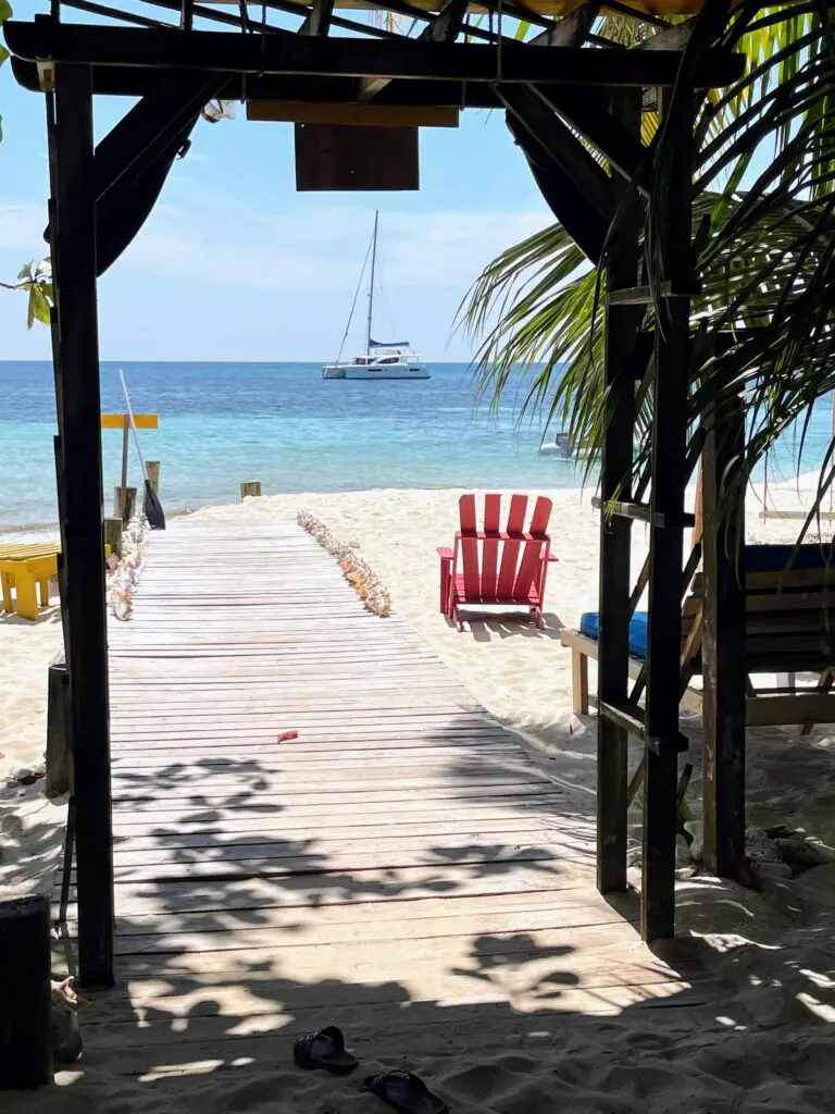 A wooden walkway leading to a beach with chairs and a sailboat.