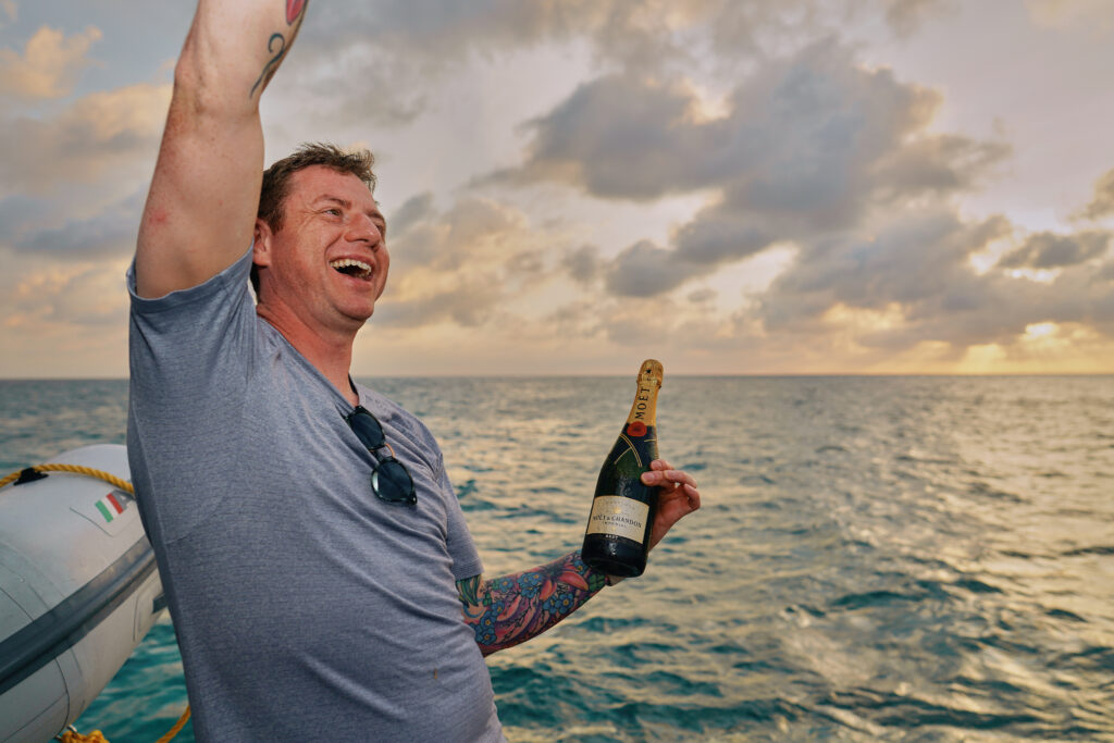 A man on a boat during his Belize sailing vacation, joyfully holding a bottle of champagne.